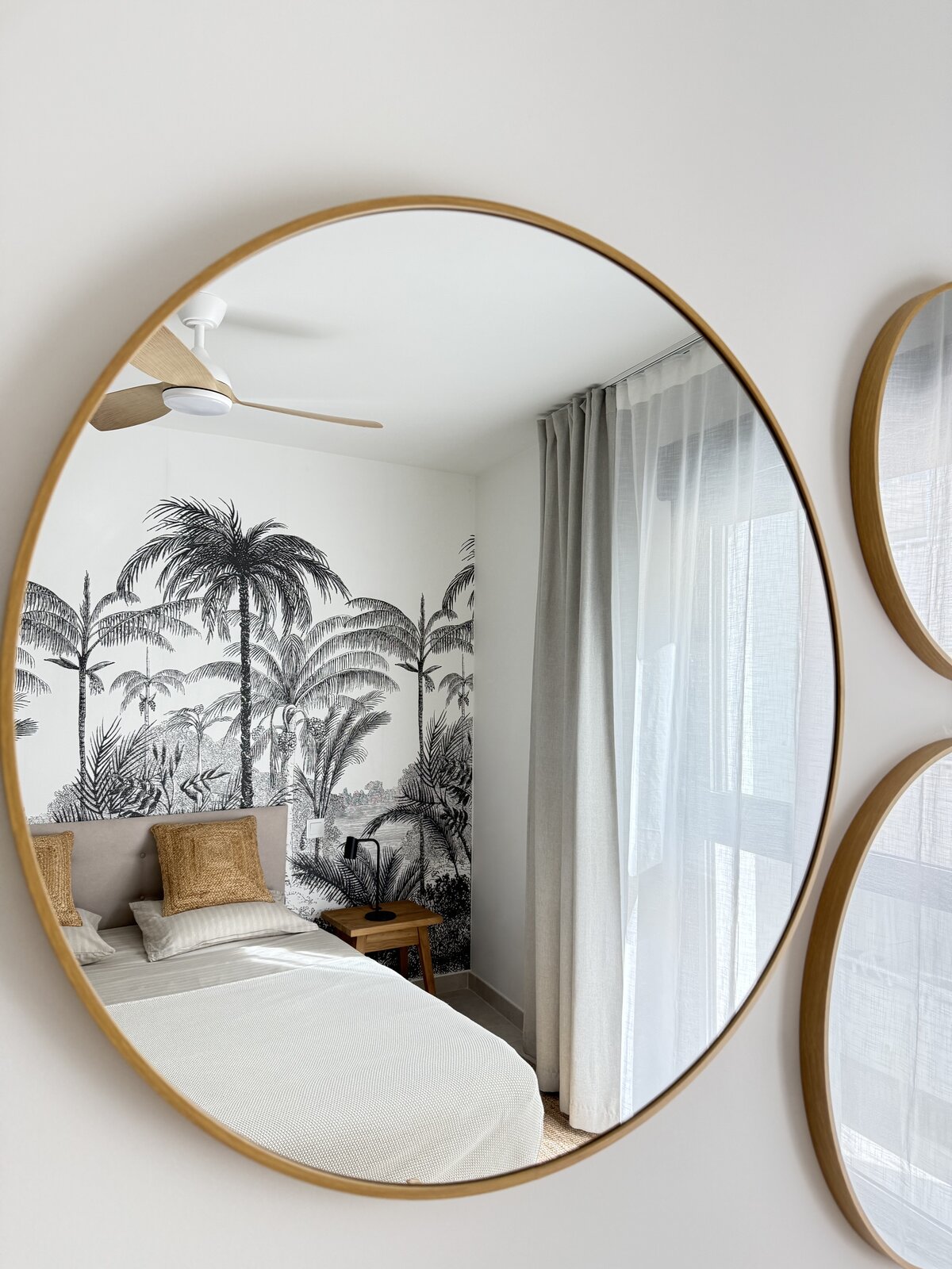 Round oak mirror reflecting bedroom with tropical palm mural and linen curtains