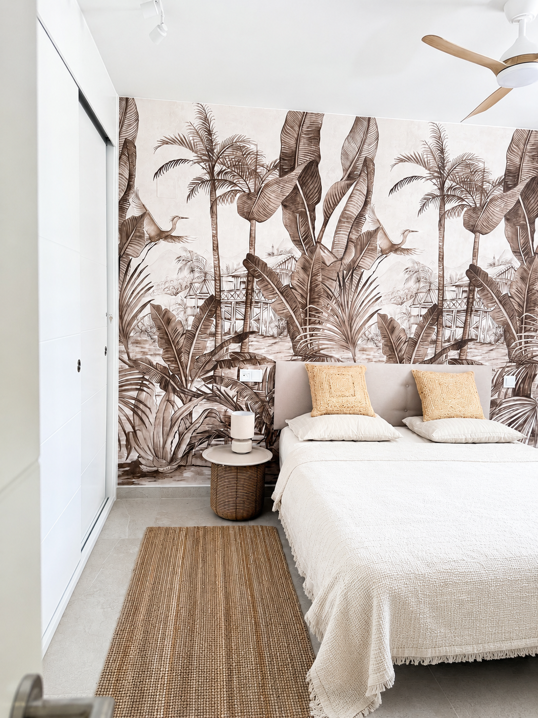 Serene bedroom with sepia tropical palm and crane mural, upholstered headboard, woven rattan side table and jute runner rug under a wooden ceiling fan