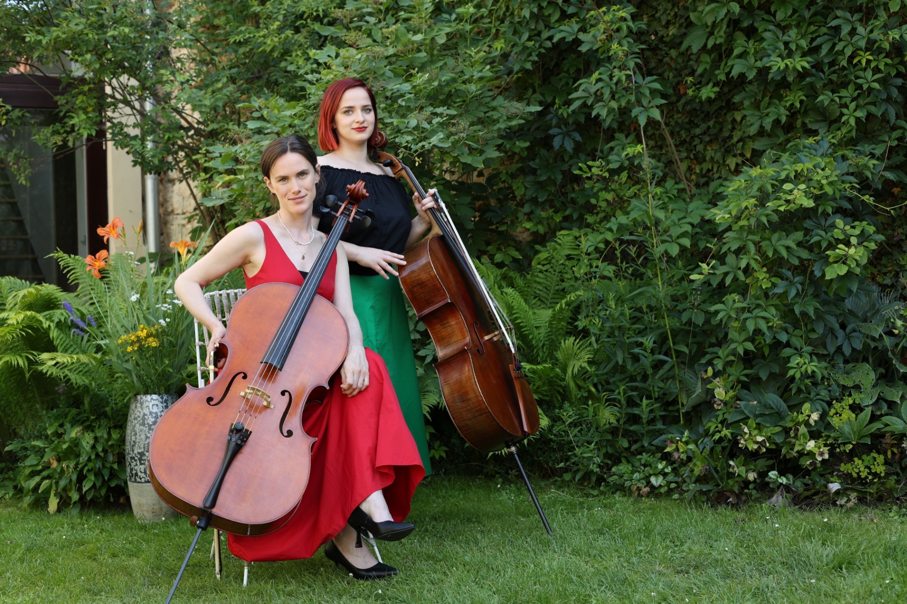 Two cellists in red and green dresses posing with their instruments in a lush summer garden
