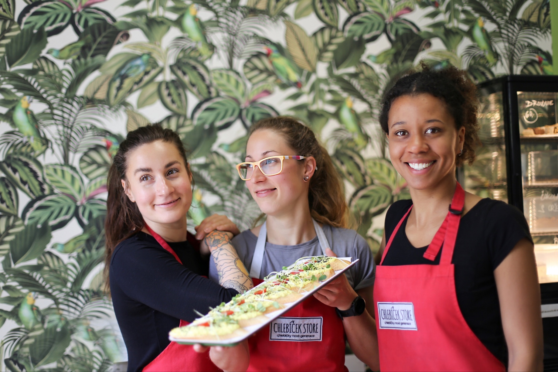 Three smiling staff members of Chlebíček Store holding a tray of open-faced sandwiches in front of tropical parrot wallpaper
