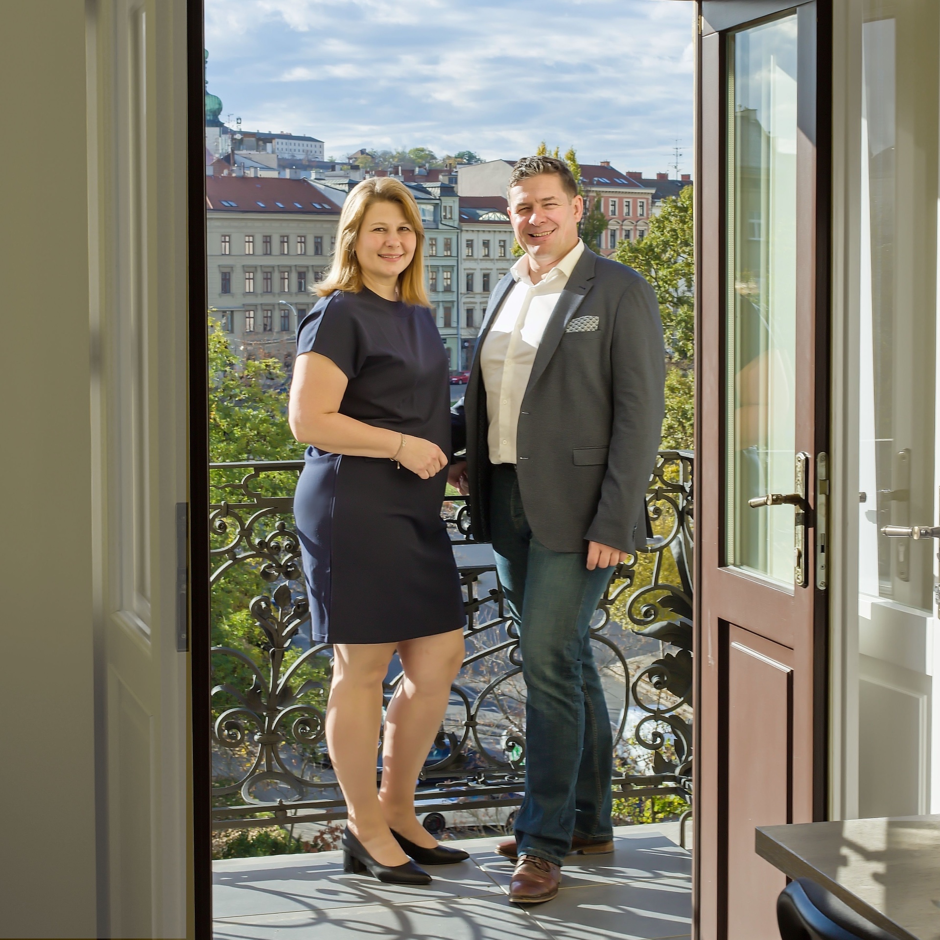 Couple standing on a wrought-iron balcony overlooking Prague rooftops, viewed through open French doors
