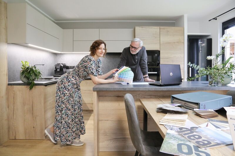 Designer in a floral dress showing colour swatches to a client at a modern kitchen island with laptop and catalogues