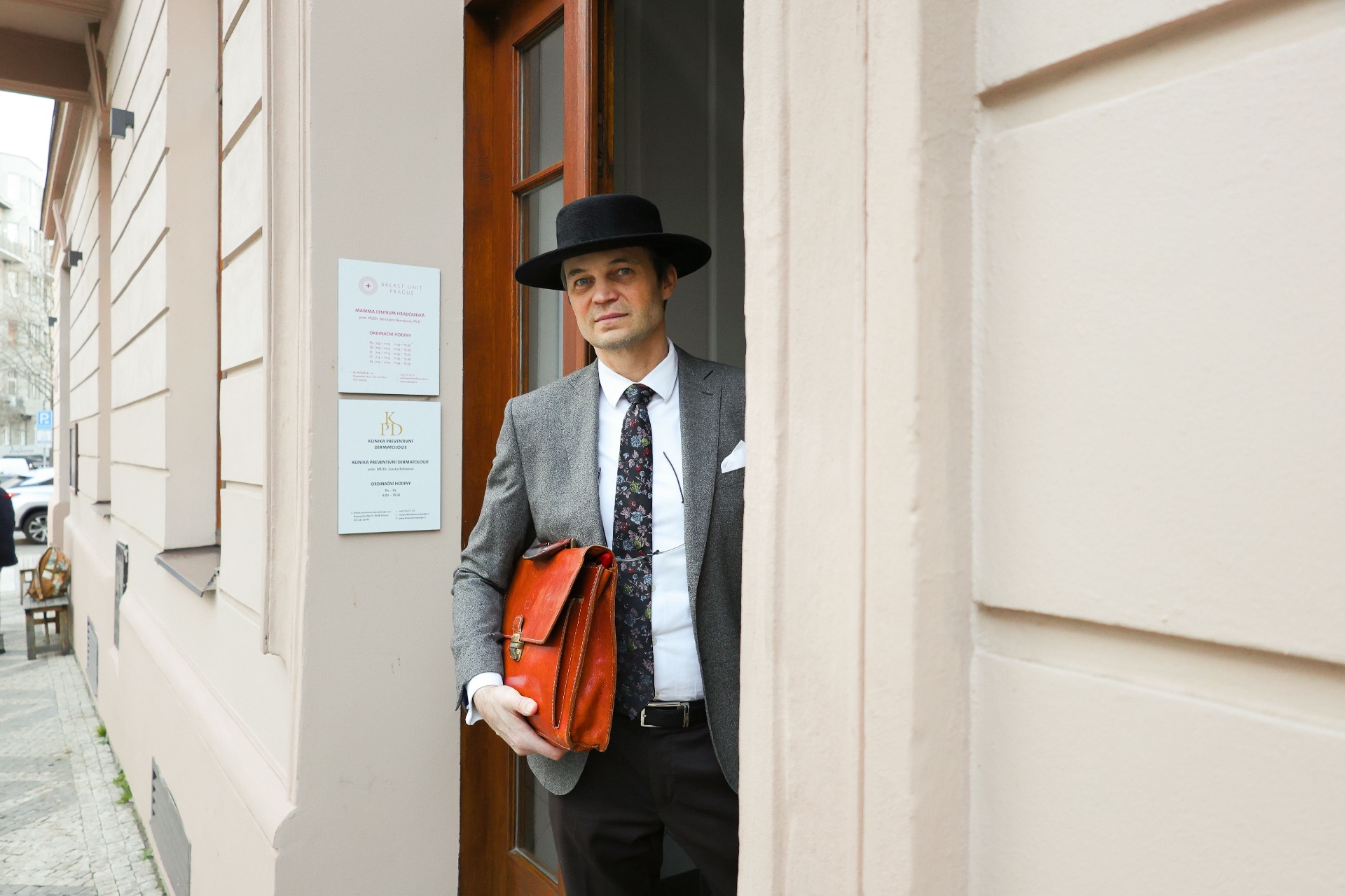 Doctor in a grey suit, black hat and red leather briefcase standing in the doorway of a Prague clinic