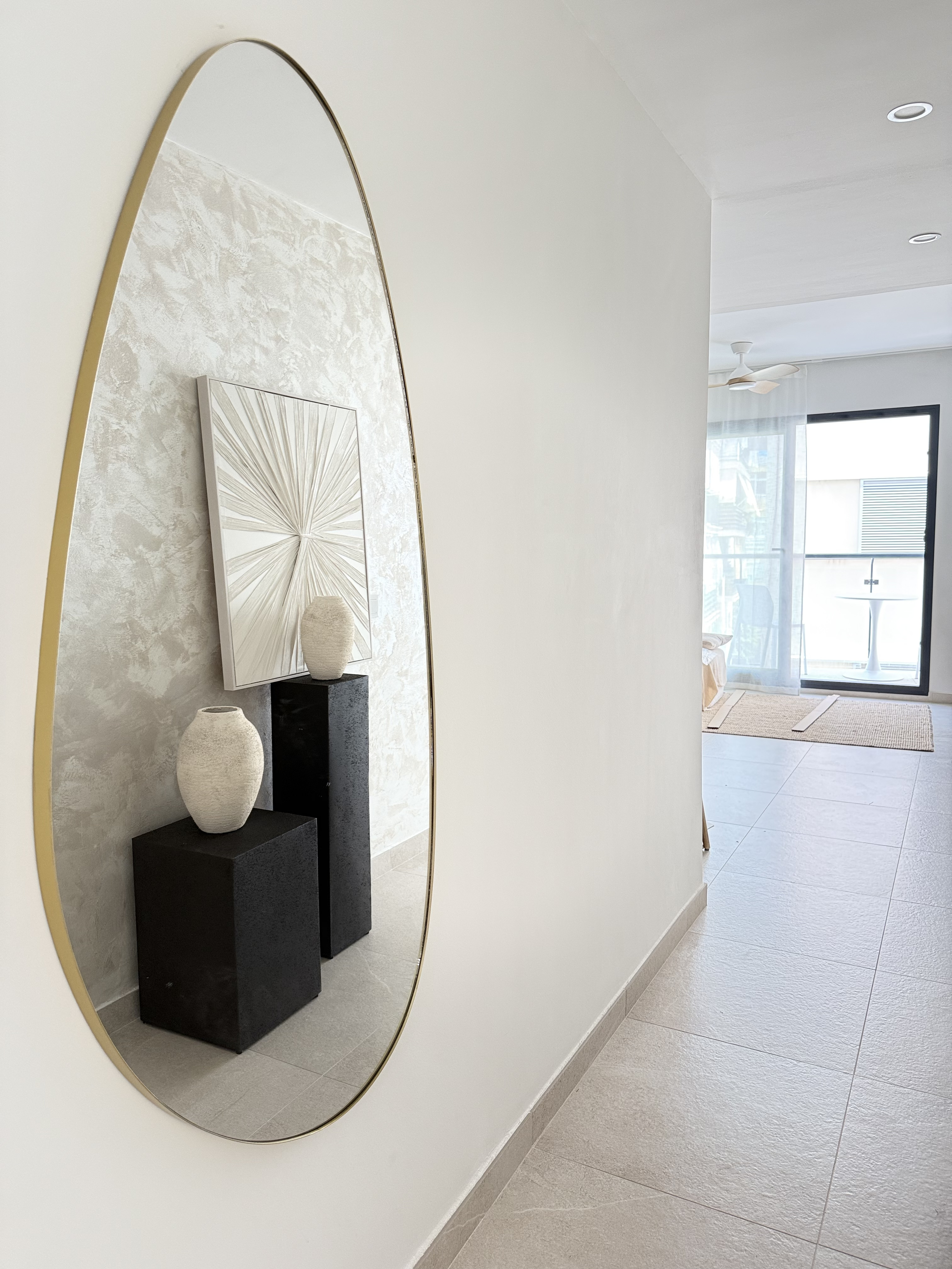 Hallway with oversized brass pebble mirror reflecting plaster wall and black plinths with ceramic vases