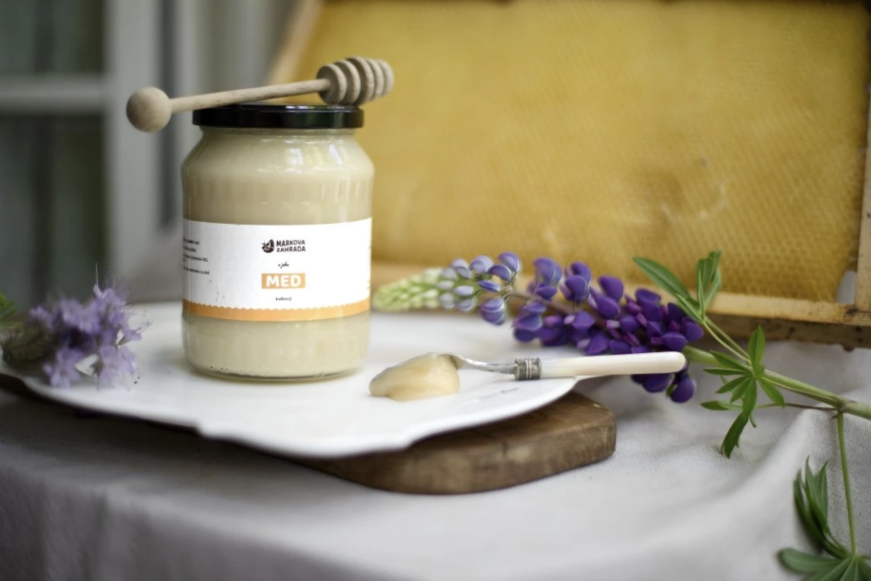 Jar of Markova Zahrada flower honey with wooden dipper, spoon and lupine on a white plate in front of a honeycomb frame