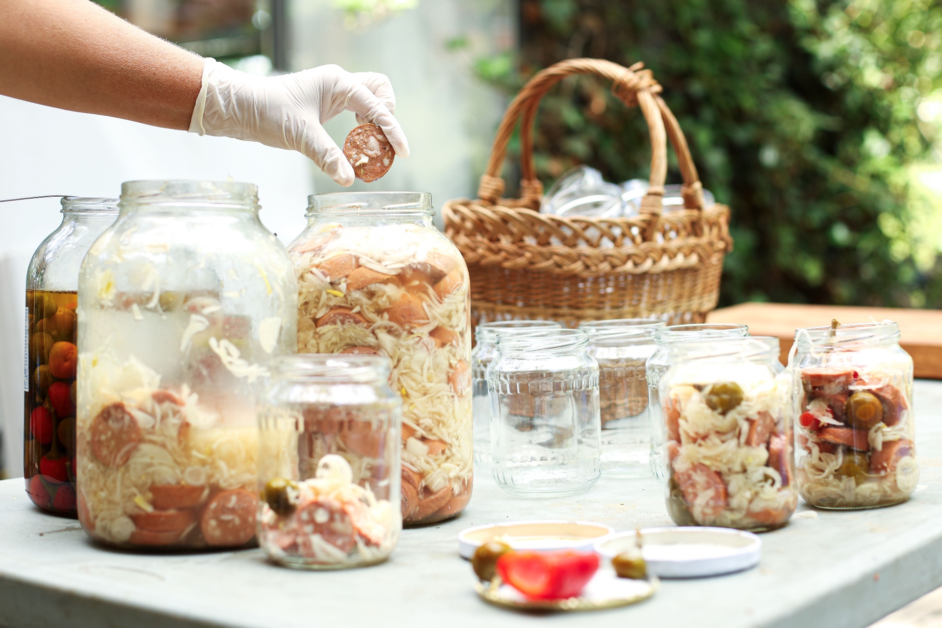 Gloved hand placing a sausage slice into a large jar of pickled sausages and onions on a garden table with wicker basket