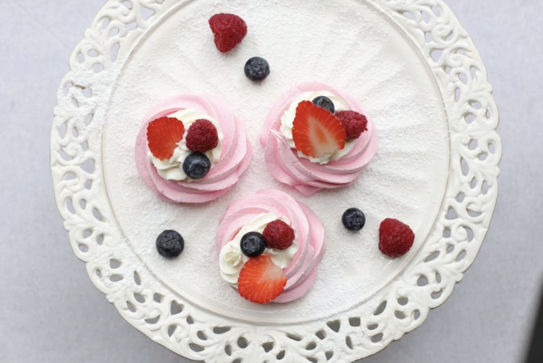 Three pink meringues topped with cream and fresh berries on a white lace cake stand