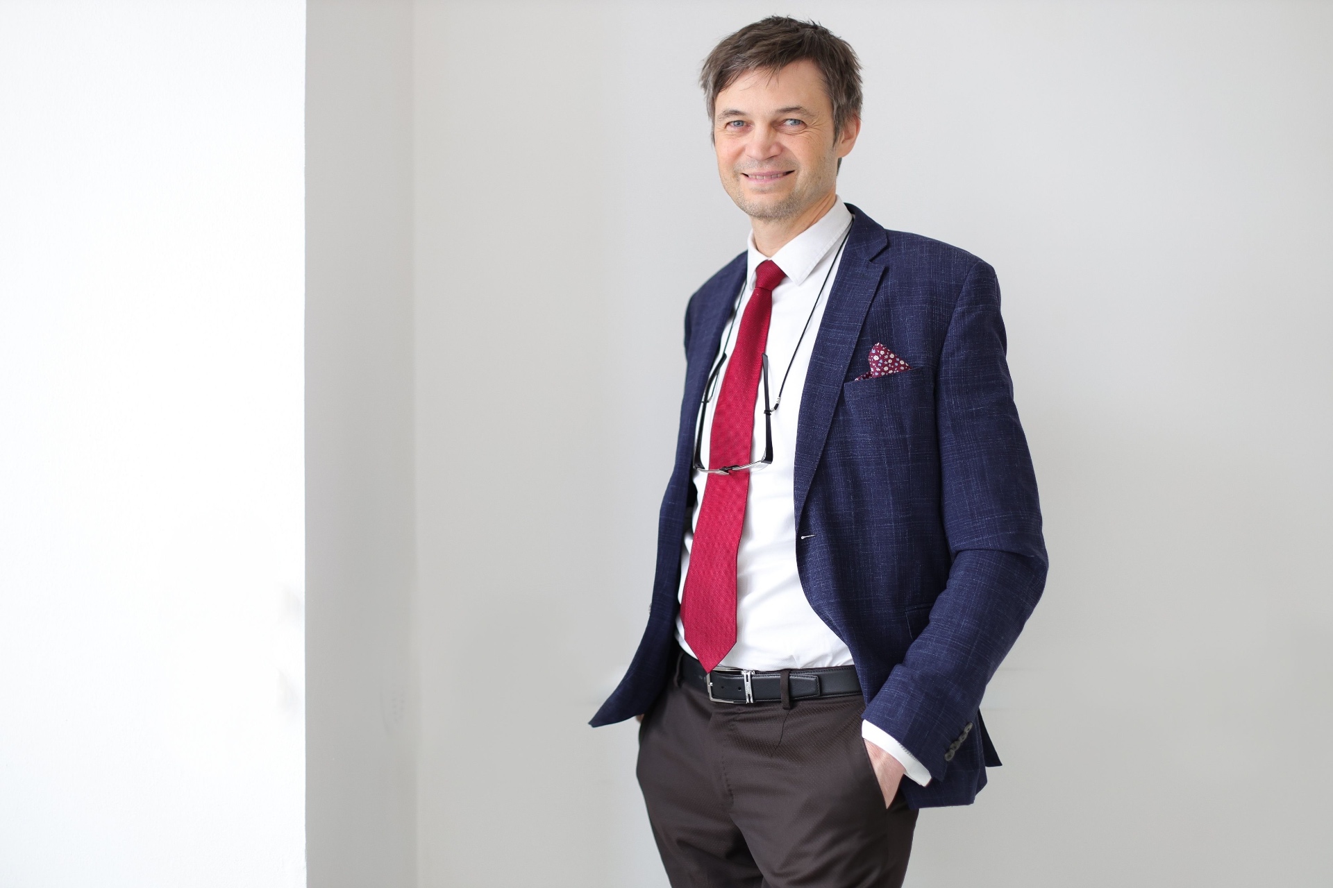 Editorial portrait of a doctor in a navy linen blazer, white shirt and red tie against a soft white wall