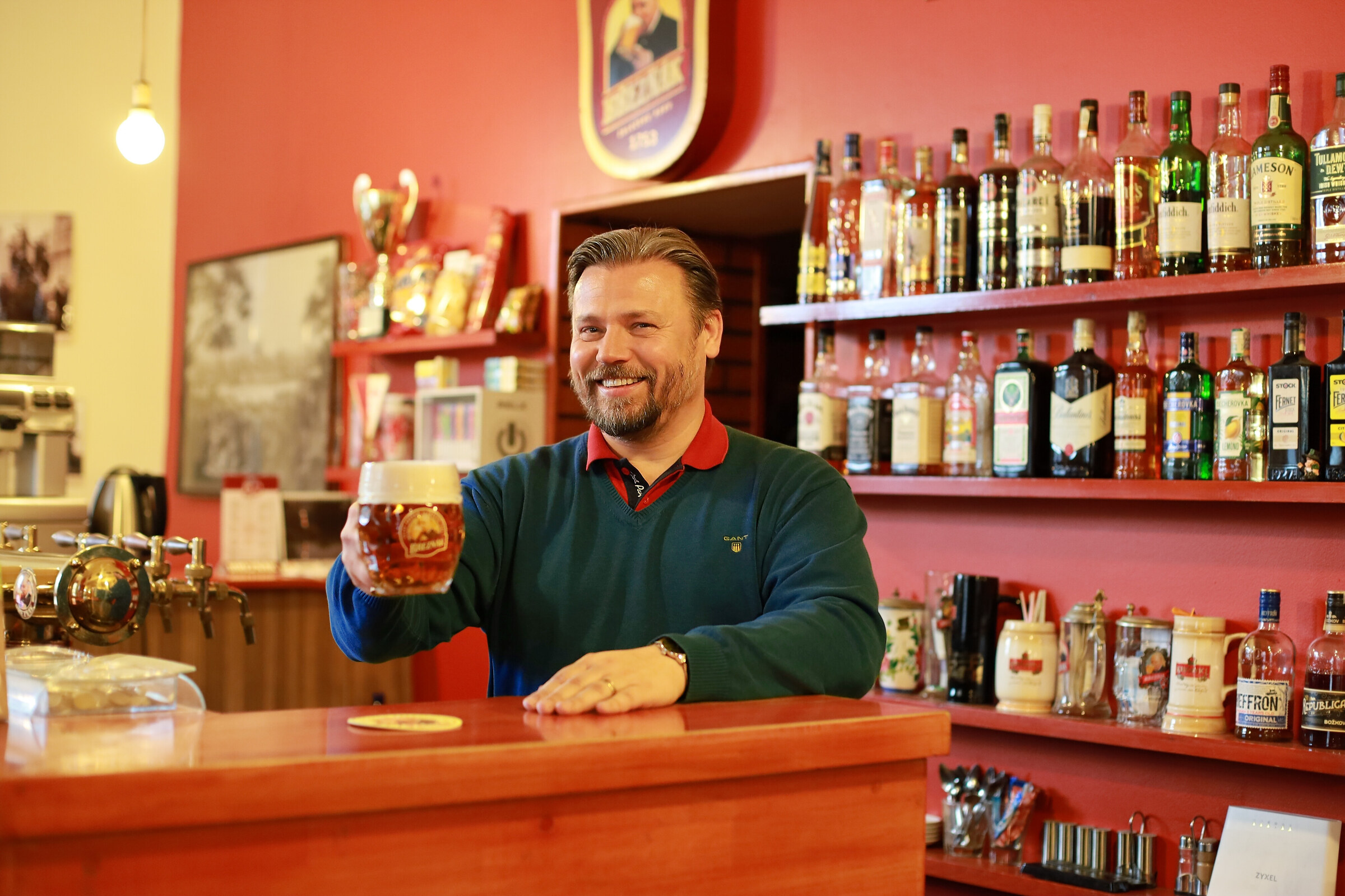 Smiling restaurateur in a green sweater raising a frothy mug of Březňák beer behind a wooden bar with red walls and shelves of spirits