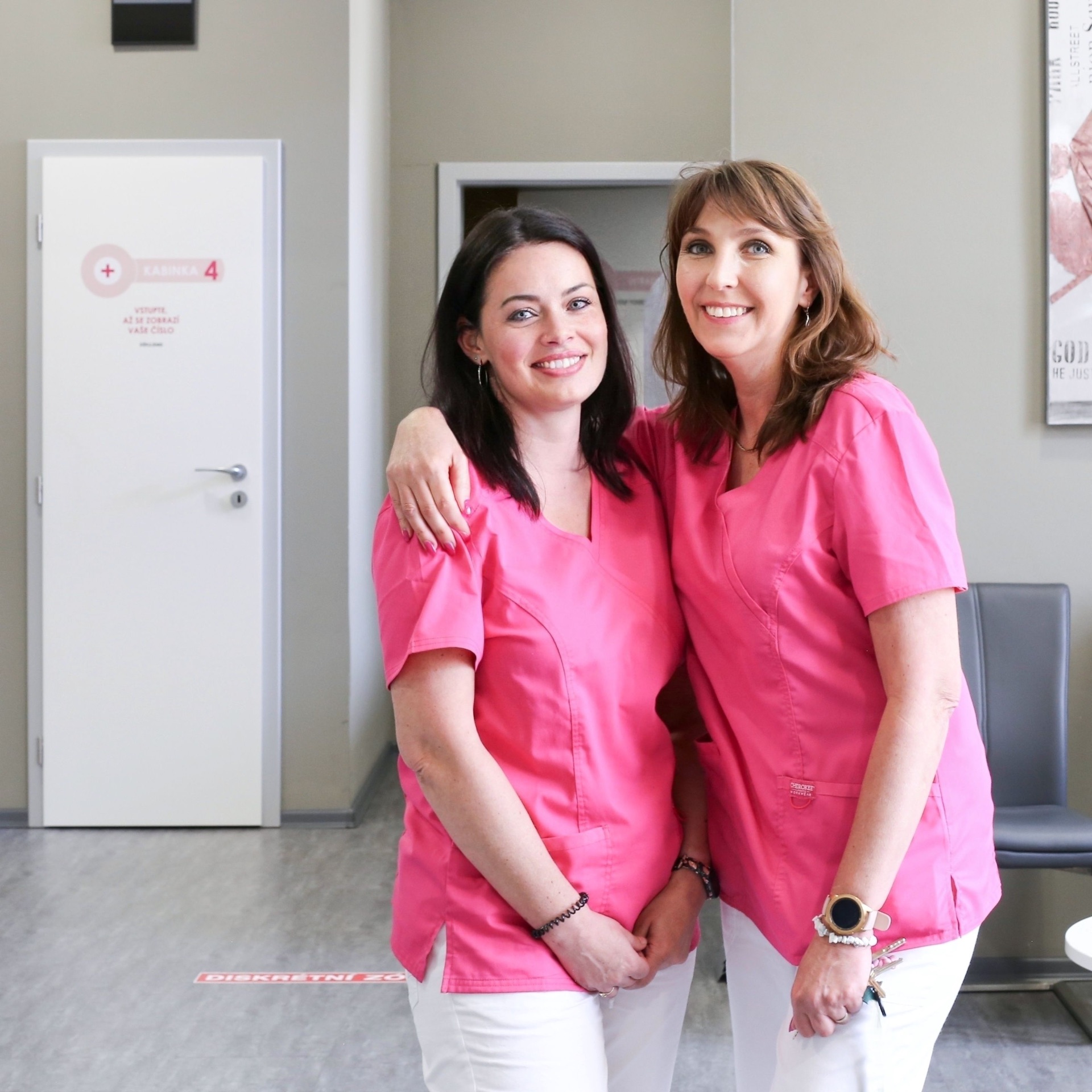 Two smiling medical assistants in pink scrubs embracing in the waiting area of a screening clinic