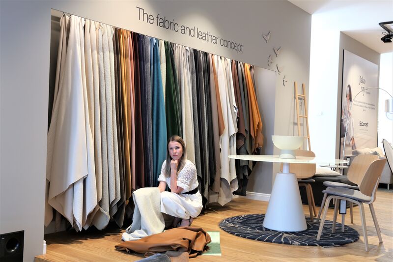Woman in a white lace dress sitting under hanging fabric samples beneath 'The fabric and leather concept' signage