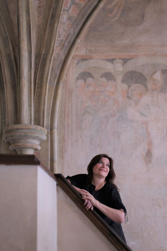 Woman in black on a wooden staircase looking up at medieval Gothic frescoes and vaulted ceiling